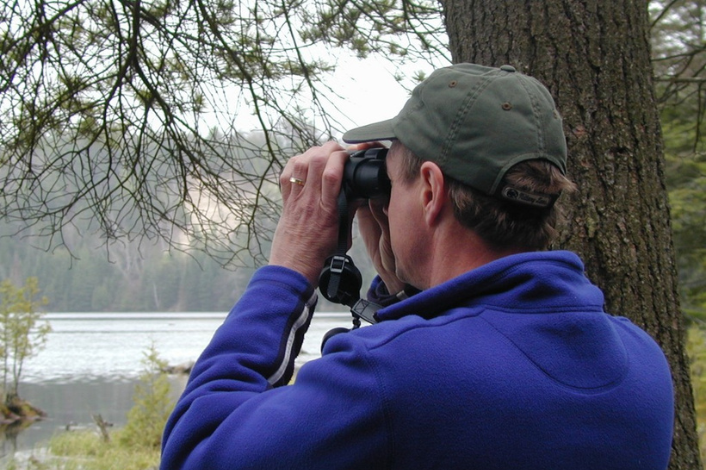 Back of a man in a blue half zip and a army green hat looking through binoculars across a body of water